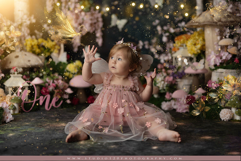 One-year-old girl celebrating her first birthday with an enchanted fairy garden cake smash, wearing a blush pink fairy dress and wings while exploring a whimsical set filled with flowers, butterflies, woodland mushrooms, lanterns, and a floral cake. Custom fairy-themed first birthday photography by Studio 120 Photography in Ramona, serving San Diego and surrounding areas.