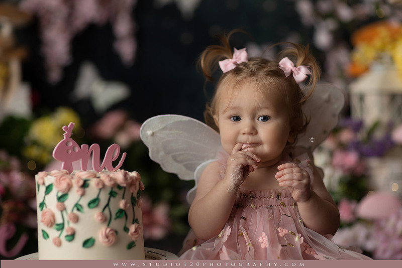 One-year-old girl celebrating her first birthday with an enchanted fairy garden cake smash, wearing a blush pink fairy dress and wings while exploring a whimsical set filled with flowers, butterflies, woodland mushrooms, lanterns, and a floral cake. Custom fairy-themed first birthday photography by Studio 120 Photography in Ramona, serving San Diego and surrounding areas.