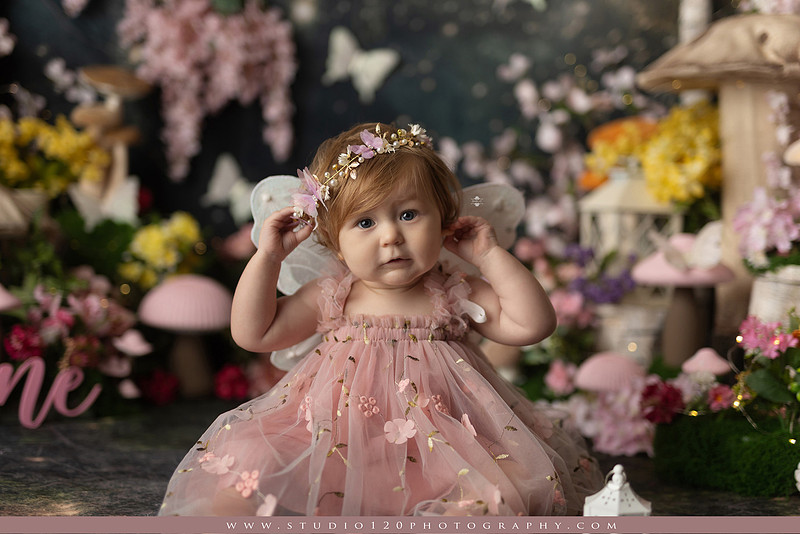 One-year-old girl celebrating her first birthday with an enchanted fairy garden cake smash, wearing a blush pink fairy dress and wings while exploring a whimsical set filled with flowers, butterflies, woodland mushrooms, lanterns, and a floral cake. Custom fairy-themed first birthday photography by Studio 120 Photography in Ramona, serving San Diego and surrounding areas.