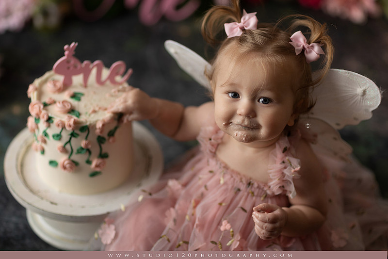 One-year-old girl celebrating her first birthday with an enchanted fairy garden cake smash, wearing a blush pink fairy dress and wings while exploring a whimsical set filled with flowers, butterflies, woodland mushrooms, lanterns, and a floral cake. Custom fairy-themed first birthday photography by Studio 120 Photography in Ramona, serving San Diego and surrounding areas.