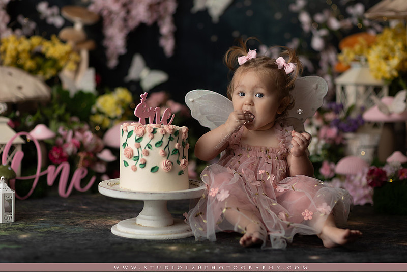 One-year-old girl celebrating her first birthday with an enchanted fairy garden cake smash, wearing a blush pink fairy dress and wings while exploring a whimsical set filled with flowers, butterflies, woodland mushrooms, lanterns, and a floral cake. Custom fairy-themed first birthday photography by Studio 120 Photography in Ramona, serving San Diego and surrounding areas.