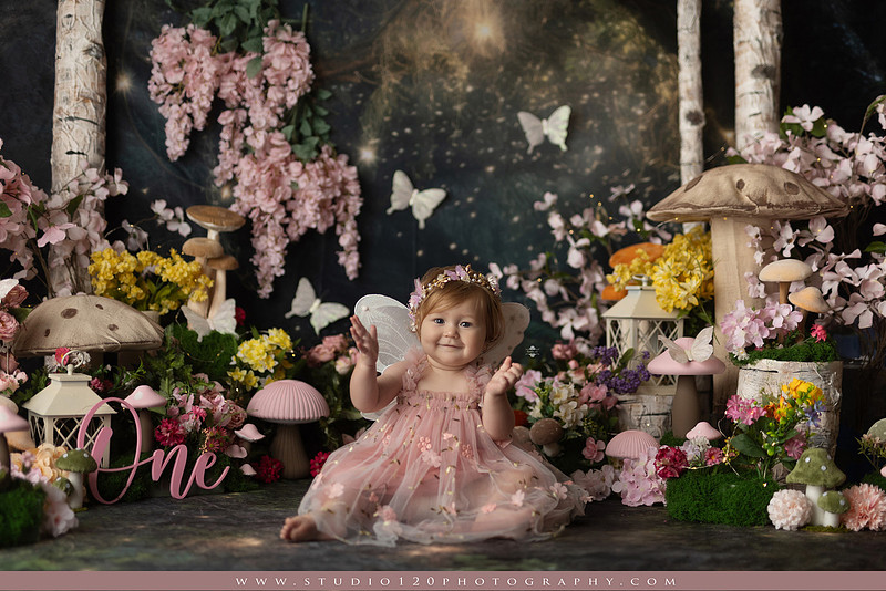 One-year-old girl celebrating her first birthday with an enchanted fairy garden cake smash, wearing a blush pink fairy dress and wings while exploring a whimsical set filled with flowers, butterflies, woodland mushrooms, lanterns, and a floral cake. Custom fairy-themed first birthday photography by Studio 120 Photography in Ramona, serving San Diego and surrounding areas.