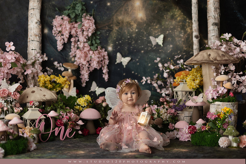 One-year-old girl celebrating her first birthday with an enchanted fairy garden cake smash, wearing a blush pink fairy dress and wings while exploring a whimsical set filled with flowers, butterflies, woodland mushrooms, lanterns, and a floral cake. Custom fairy-themed first birthday photography by Studio 120 Photography in Ramona, serving San Diego and surrounding areas.