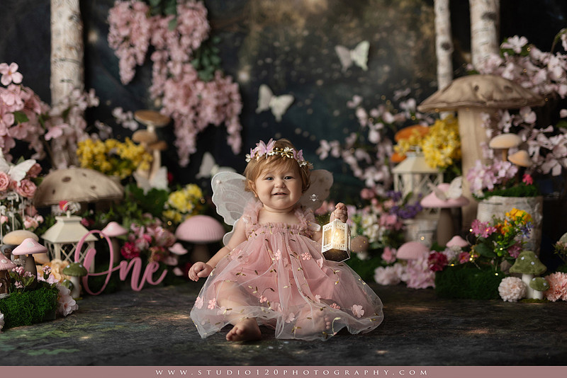 One-year-old girl celebrating her first birthday with an enchanted fairy garden cake smash, wearing a blush pink fairy dress and wings while exploring a whimsical set filled with flowers, butterflies, woodland mushrooms, lanterns, and a floral cake. Custom fairy-themed first birthday photography by Studio 120 Photography in Ramona, serving San Diego and surrounding areas.