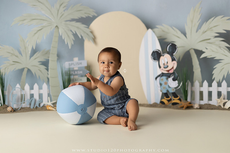 A beach-themed Mickey Mouse first birthday cake smash featuring a smiling one-year-old boy in a custom coastal set with palm trees, surfboards, seashells, beach signs, a white picket fence, and soft blue ocean-inspired décor. The gallery includes clean milestone portraits in a denim romper before the cake, followed by joyful cake smash photos with a blue and white birthday cake, frosting-covered smiles, playful expressions, and happy moments celebrating a first birthday. Captured at Studio 120 Photography in Ramona, serving families throughout San Diego with custom cake smash and milestone photography.