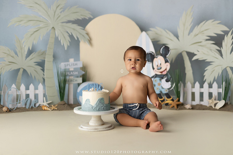 A beach-themed Mickey Mouse first birthday cake smash featuring a smiling one-year-old boy in a custom coastal set with palm trees, surfboards, seashells, beach signs, a white picket fence, and soft blue ocean-inspired décor. The gallery includes clean milestone portraits in a denim romper before the cake, followed by joyful cake smash photos with a blue and white birthday cake, frosting-covered smiles, playful expressions, and happy moments celebrating a first birthday. Captured at Studio 120 Photography in Ramona, serving families throughout San Diego with custom cake smash and milestone photography.