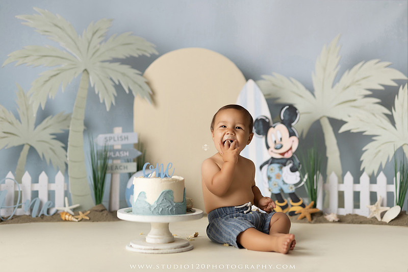 A beach-themed Mickey Mouse first birthday cake smash featuring a smiling one-year-old boy in a custom coastal set with palm trees, surfboards, seashells, beach signs, a white picket fence, and soft blue ocean-inspired décor. The gallery includes clean milestone portraits in a denim romper before the cake, followed by joyful cake smash photos with a blue and white birthday cake, frosting-covered smiles, playful expressions, and happy moments celebrating a first birthday. Captured at Studio 120 Photography in Ramona, serving families throughout San Diego with custom cake smash and milestone photography.