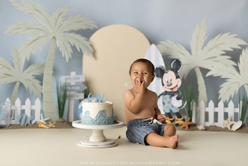 A beach-themed Mickey Mouse first birthday cake smash featuring a smiling one-year-old boy in a custom coastal set with palm trees, surfboards, seashells, beach signs, a white picket fence, and soft blue ocean-inspired décor. The gallery includes clean milestone portraits in a denim romper before the cake, followed by joyful cake smash photos with a blue and white birthday cake, frosting-covered smiles, playful expressions, and happy moments celebrating a first birthday. Captured at Studio 120 Photography in Ramona, serving families throughout San Diego with custom cake smash and milestone photography.