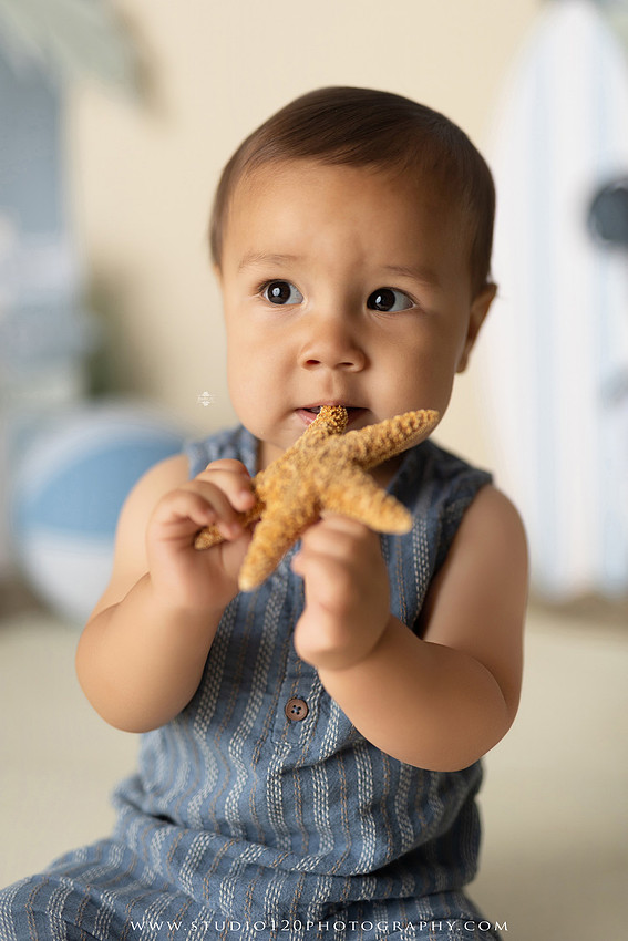 A beach-themed Mickey Mouse first birthday cake smash featuring a smiling one-year-old boy in a custom coastal set with palm trees, surfboards, seashells, beach signs, a white picket fence, and soft blue ocean-inspired décor. The gallery includes clean milestone portraits in a denim romper before the cake, followed by joyful cake smash photos with a blue and white birthday cake, frosting-covered smiles, playful expressions, and happy moments celebrating a first birthday. Captured at Studio 120 Photography in Ramona, serving families throughout San Diego with custom cake smash and milestone photography.