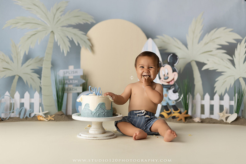 A beach-themed Mickey Mouse first birthday cake smash featuring a smiling one-year-old boy in a custom coastal set with palm trees, surfboards, seashells, beach signs, a white picket fence, and soft blue ocean-inspired décor. The gallery includes clean milestone portraits in a denim romper before the cake, followed by joyful cake smash photos with a blue and white birthday cake, frosting-covered smiles, playful expressions, and happy moments celebrating a first birthday. Captured at Studio 120 Photography in Ramona, serving families throughout San Diego with custom cake smash and milestone photography.
