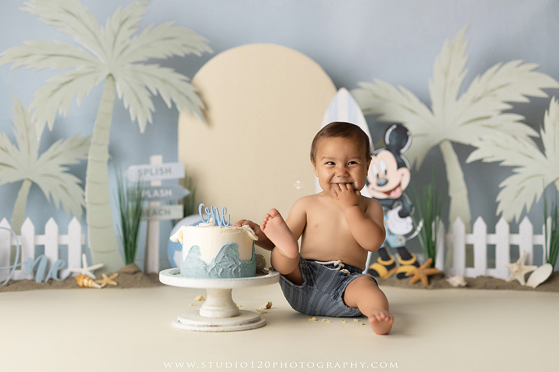 A beach-themed Mickey Mouse first birthday cake smash featuring a smiling one-year-old boy in a custom coastal set with palm trees, surfboards, seashells, beach signs, a white picket fence, and soft blue ocean-inspired décor. The gallery includes clean milestone portraits in a denim romper before the cake, followed by joyful cake smash photos with a blue and white birthday cake, frosting-covered smiles, playful expressions, and happy moments celebrating a first birthday. Captured at Studio 120 Photography in Ramona, serving families throughout San Diego with custom cake smash and milestone photography.