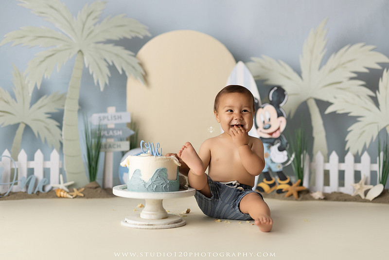 A beach-themed Mickey Mouse first birthday cake smash featuring a smiling one-year-old boy in a custom coastal set with palm trees, surfboards, seashells, beach signs, a white picket fence, and soft blue ocean-inspired décor. The gallery includes clean milestone portraits in a denim romper before the cake, followed by joyful cake smash photos with a blue and white birthday cake, frosting-covered smiles, playful expressions, and happy moments celebrating a first birthday. Captured at Studio 120 Photography in Ramona, serving families throughout San Diego with custom cake smash and milestone photography.