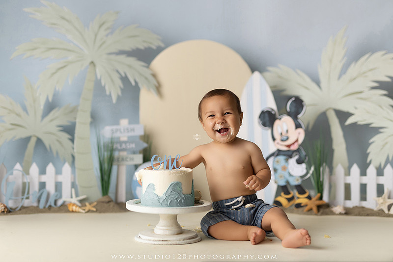 A beach-themed Mickey Mouse first birthday cake smash featuring a smiling one-year-old boy in a custom coastal set with palm trees, surfboards, seashells, beach signs, a white picket fence, and soft blue ocean-inspired décor. The gallery includes clean milestone portraits in a denim romper before the cake, followed by joyful cake smash photos with a blue and white birthday cake, frosting-covered smiles, playful expressions, and happy moments celebrating a first birthday. Captured at Studio 120 Photography in Ramona, serving families throughout San Diego with custom cake smash and milestone photography.