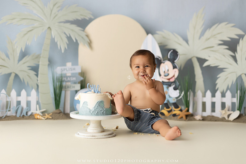 A beach-themed Mickey Mouse first birthday cake smash featuring a smiling one-year-old boy in a custom coastal set with palm trees, surfboards, seashells, beach signs, a white picket fence, and soft blue ocean-inspired décor. The gallery includes clean milestone portraits in a denim romper before the cake, followed by joyful cake smash photos with a blue and white birthday cake, frosting-covered smiles, playful expressions, and happy moments celebrating a first birthday. Captured at Studio 120 Photography in Ramona, serving families throughout San Diego with custom cake smash and milestone photography.