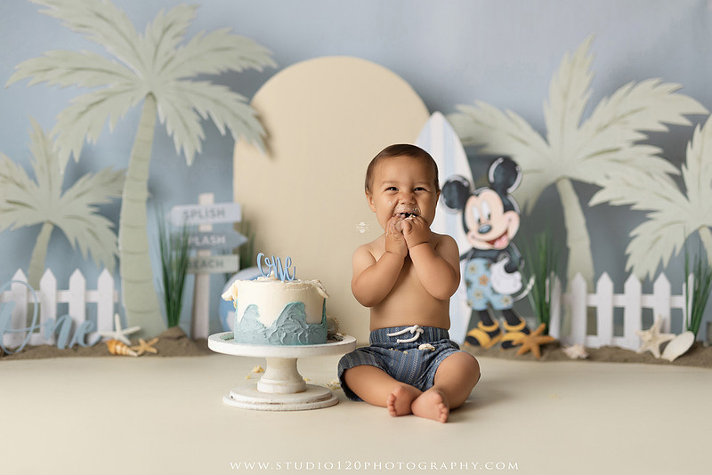 A beach-themed Mickey Mouse first birthday cake smash featuring a smiling one-year-old boy in a custom coastal set with palm trees, surfboards, seashells, beach signs, a white picket fence, and soft blue ocean-inspired décor. The gallery includes clean milestone portraits in a denim romper before the cake, followed by joyful cake smash photos with a blue and white birthday cake, frosting-covered smiles, playful expressions, and happy moments celebrating a first birthday. Captured at Studio 120 Photography in Ramona, serving families throughout San Diego with custom cake smash and milestone photography.