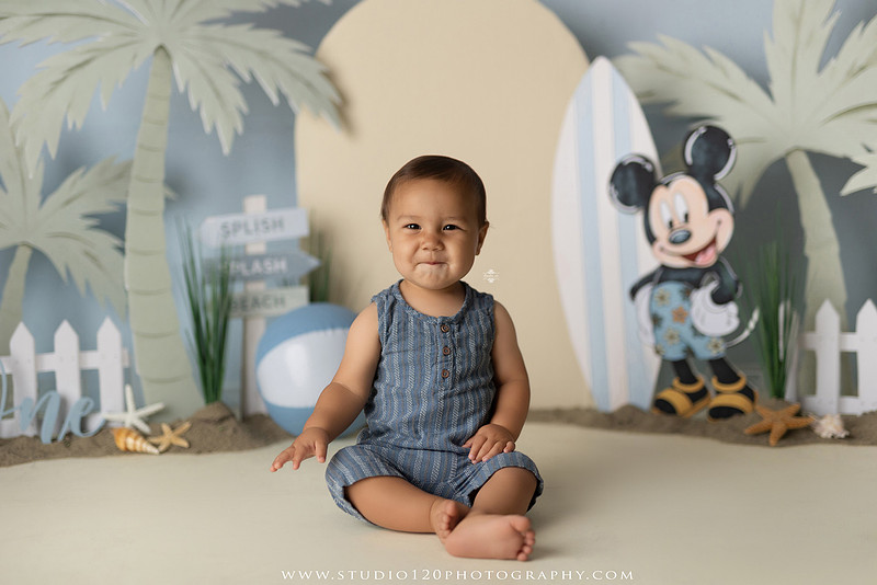 A beach-themed Mickey Mouse first birthday cake smash featuring a smiling one-year-old boy in a custom coastal set with palm trees, surfboards, seashells, beach signs, a white picket fence, and soft blue ocean-inspired décor. The gallery includes clean milestone portraits in a denim romper before the cake, followed by joyful cake smash photos with a blue and white birthday cake, frosting-covered smiles, playful expressions, and happy moments celebrating a first birthday. Captured at Studio 120 Photography in Ramona, serving families throughout San Diego with custom cake smash and milestone photography.