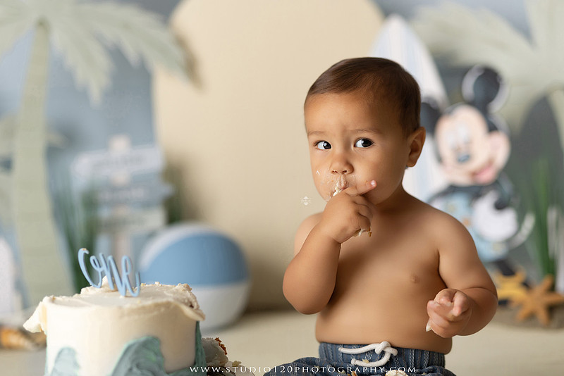 A beach-themed Mickey Mouse first birthday cake smash featuring a smiling one-year-old boy in a custom coastal set with palm trees, surfboards, seashells, beach signs, a white picket fence, and soft blue ocean-inspired décor. The gallery includes clean milestone portraits in a denim romper before the cake, followed by joyful cake smash photos with a blue and white birthday cake, frosting-covered smiles, playful expressions, and happy moments celebrating a first birthday. Captured at Studio 120 Photography in Ramona, serving families throughout San Diego with custom cake smash and milestone photography.