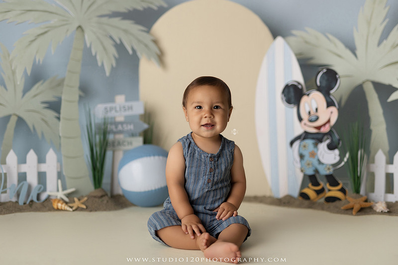 A beach-themed Mickey Mouse first birthday cake smash featuring a smiling one-year-old boy in a custom coastal set with palm trees, surfboards, seashells, beach signs, a white picket fence, and soft blue ocean-inspired décor. The gallery includes clean milestone portraits in a denim romper before the cake, followed by joyful cake smash photos with a blue and white birthday cake, frosting-covered smiles, playful expressions, and happy moments celebrating a first birthday. Captured at Studio 120 Photography in Ramona, serving families throughout San Diego with custom cake smash and milestone photography.
