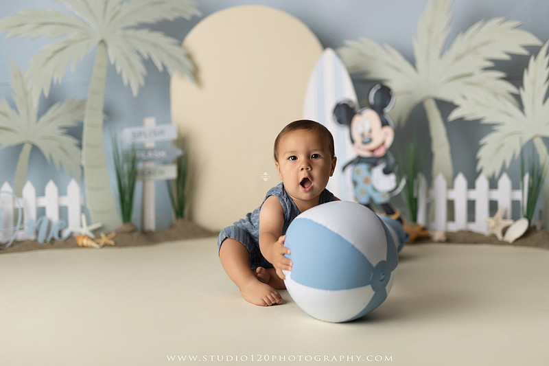 A beach-themed Mickey Mouse first birthday cake smash featuring a smiling one-year-old boy in a custom coastal set with palm trees, surfboards, seashells, beach signs, a white picket fence, and soft blue ocean-inspired décor. The gallery includes clean milestone portraits in a denim romper before the cake, followed by joyful cake smash photos with a blue and white birthday cake, frosting-covered smiles, playful expressions, and happy moments celebrating a first birthday. Captured at Studio 120 Photography in Ramona, serving families throughout San Diego with custom cake smash and milestone photography.
