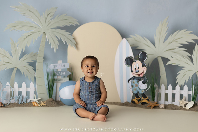 A beach-themed Mickey Mouse first birthday cake smash featuring a smiling one-year-old boy in a custom coastal set with palm trees, surfboards, seashells, beach signs, a white picket fence, and soft blue ocean-inspired décor. The gallery includes clean milestone portraits in a denim romper before the cake, followed by joyful cake smash photos with a blue and white birthday cake, frosting-covered smiles, playful expressions, and happy moments celebrating a first birthday. Captured at Studio 120 Photography in Ramona, serving families throughout San Diego with custom cake smash and milestone photography.