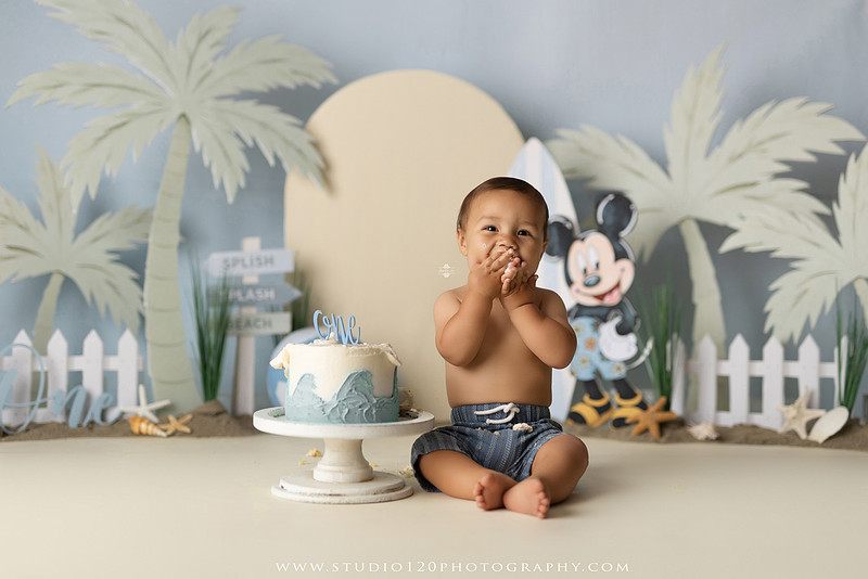 A beach-themed Mickey Mouse first birthday cake smash featuring a smiling one-year-old boy in a custom coastal set with palm trees, surfboards, seashells, beach signs, a white picket fence, and soft blue ocean-inspired décor. The gallery includes clean milestone portraits in a denim romper before the cake, followed by joyful cake smash photos with a blue and white birthday cake, frosting-covered smiles, playful expressions, and happy moments celebrating a first birthday. Captured at Studio 120 Photography in Ramona, serving families throughout San Diego with custom cake smash and milestone photography.