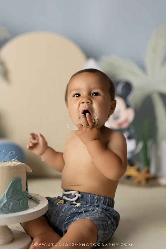 A beach-themed Mickey Mouse first birthday cake smash featuring a smiling one-year-old boy in a custom coastal set with palm trees, surfboards, seashells, beach signs, a white picket fence, and soft blue ocean-inspired décor. The gallery includes clean milestone portraits in a denim romper before the cake, followed by joyful cake smash photos with a blue and white birthday cake, frosting-covered smiles, playful expressions, and happy moments celebrating a first birthday. Captured at Studio 120 Photography in Ramona, serving families throughout San Diego with custom cake smash and milestone photography.