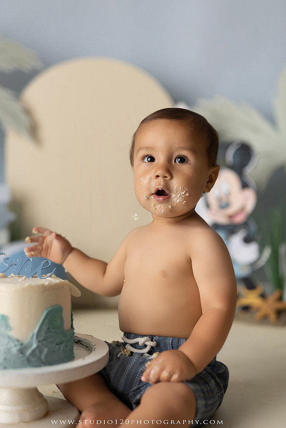 A beach-themed Mickey Mouse first birthday cake smash featuring a smiling one-year-old boy in a custom coastal set with palm trees, surfboards, seashells, beach signs, a white picket fence, and soft blue ocean-inspired décor. The gallery includes clean milestone portraits in a denim romper before the cake, followed by joyful cake smash photos with a blue and white birthday cake, frosting-covered smiles, playful expressions, and happy moments celebrating a first birthday. Captured at Studio 120 Photography in Ramona, serving families throughout San Diego with custom cake smash and milestone photography.
