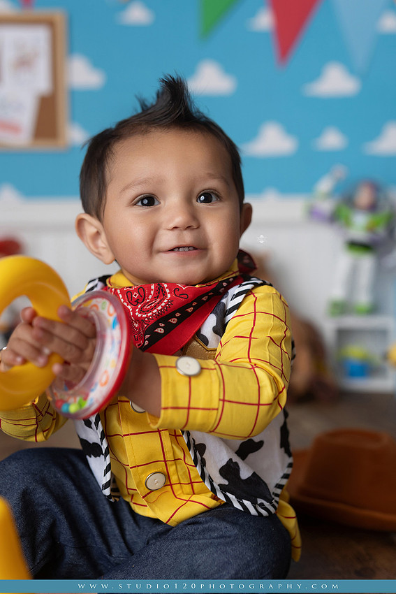 One-year-old boy dressed as Woody from Toy Story during a colorful milestone photography session, playing with classic toys including Rex the dinosaur, a cowboy hat, and ring toss games on a custom Toy Story inspired first birthday set at Studio 120 Photography in Ramona, serving San Diego and surrounding areas.