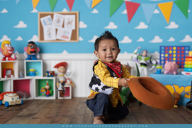 One-year-old boy dressed as Woody from Toy Story during a colorful milestone photography session, playing with classic toys including Rex the dinosaur, a cowboy hat, and ring toss games on a custom Toy Story inspired first birthday set at Studio 120 Photography in Ramona, serving San Diego and surrounding areas.