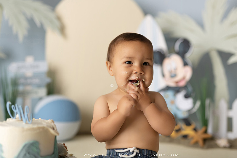 A beach-themed Mickey Mouse first birthday cake smash featuring a smiling one-year-old boy in a custom coastal set with palm trees, surfboards, seashells, beach signs, a white picket fence, and soft blue ocean-inspired décor. The gallery includes clean milestone portraits in a denim romper before the cake, followed by joyful cake smash photos with a blue and white birthday cake, frosting-covered smiles, playful expressions, and happy moments celebrating a first birthday. Captured at Studio 120 Photography in Ramona, serving families throughout San Diego with custom cake smash and milestone photography.