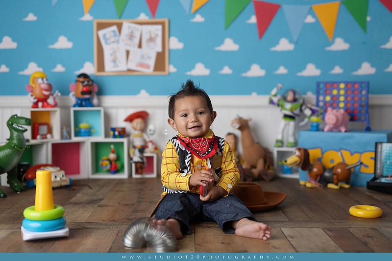 One-year-old boy dressed as Woody from Toy Story during a colorful milestone photography session, playing with classic toys including Rex the dinosaur, a cowboy hat, and ring toss games on a custom Toy Story inspired first birthday set at Studio 120 Photography in Ramona, serving San Diego and surrounding areas.
