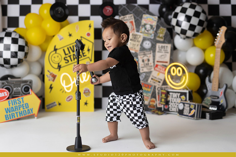 One-year-old boy enjoying a Vans Warped Tour inspired first birthday milestone photography session, posing with a microphone, guitar, concert backdrop, checkerboard details, balloons, and rock concert props on a custom music-themed set at Studio 120 Photography in Ramona, serving San Diego and surrounding areas.