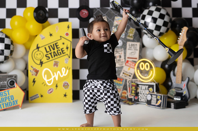 One-year-old boy enjoying a Vans Warped Tour inspired first birthday milestone photography session, posing with a microphone, guitar, concert backdrop, checkerboard details, balloons, and rock concert props on a custom music-themed set at Studio 120 Photography in Ramona, serving San Diego and surrounding areas.
