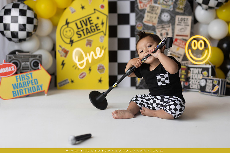 One-year-old boy enjoying a Vans Warped Tour inspired first birthday milestone photography session, posing with a microphone, guitar, concert backdrop, checkerboard details, balloons, and rock concert props on a custom music-themed set at Studio 120 Photography in Ramona, serving San Diego and surrounding areas.