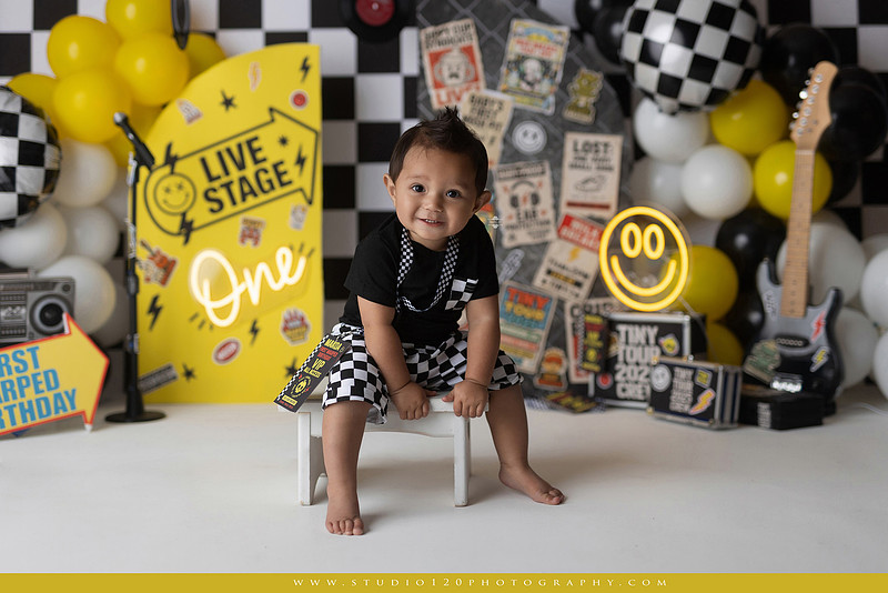 One-year-old boy enjoying a Vans Warped Tour inspired first birthday milestone photography session, posing with a microphone, guitar, concert backdrop, checkerboard details, balloons, and rock concert props on a custom music-themed set at Studio 120 Photography in Ramona, serving San Diego and surrounding areas.