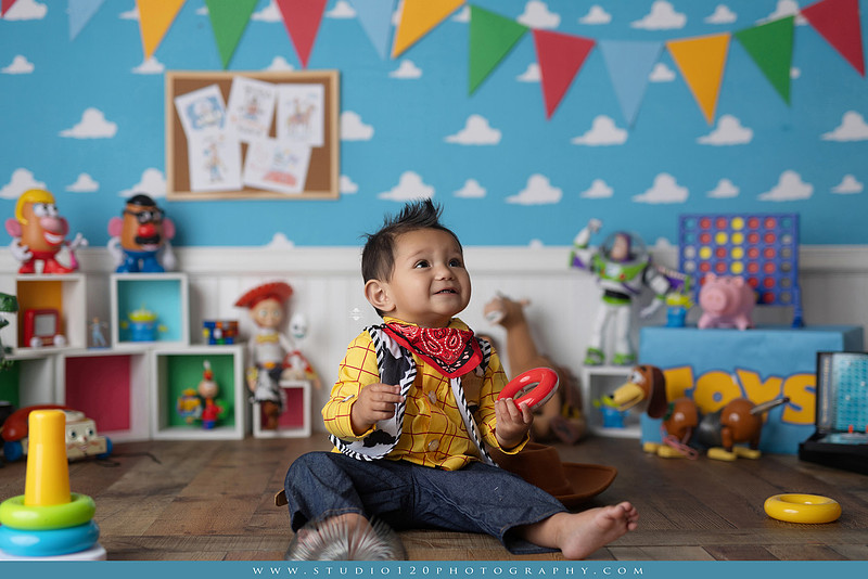 One-year-old boy dressed as Woody from Toy Story during a colorful milestone photography session, playing with classic toys including Rex the dinosaur, a cowboy hat, and ring toss games on a custom Toy Story inspired first birthday set at Studio 120 Photography in Ramona, serving San Diego and surrounding areas.
