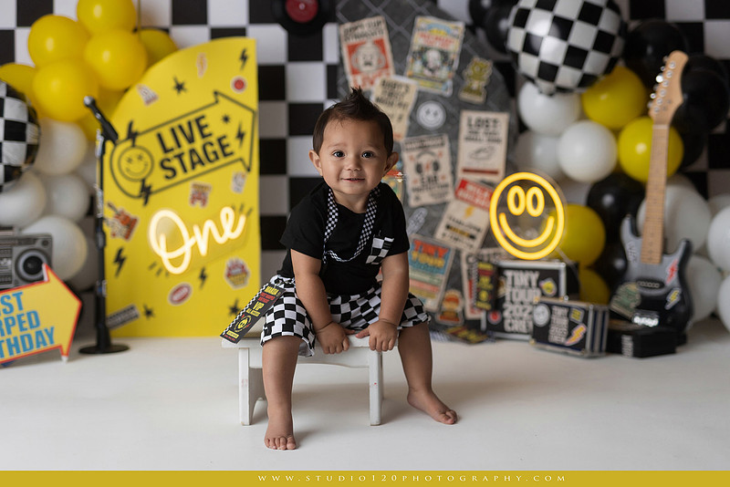 One-year-old boy enjoying a Vans Warped Tour inspired first birthday milestone photography session, posing with a microphone, guitar, concert backdrop, checkerboard details, balloons, and rock concert props on a custom music-themed set at Studio 120 Photography in Ramona, serving San Diego and surrounding areas.
