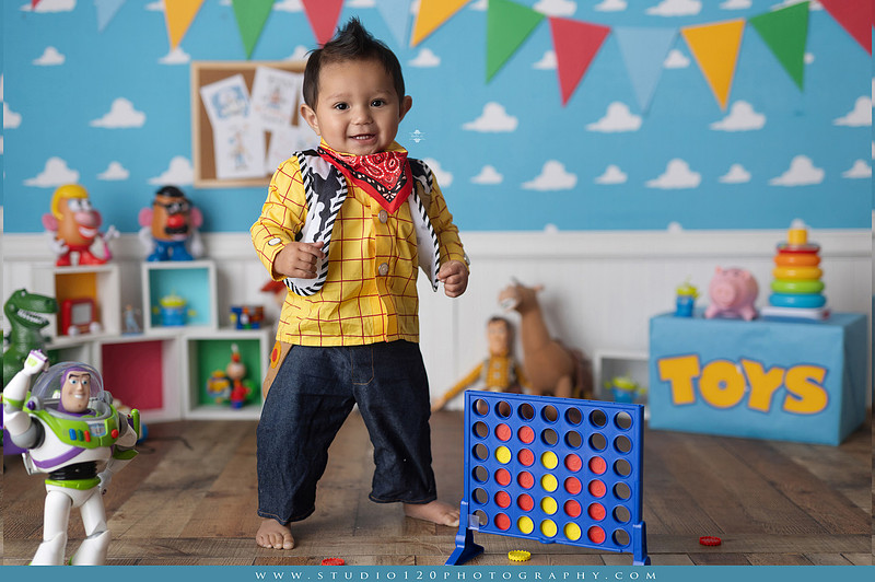 One-year-old boy dressed as Woody from Toy Story during a colorful milestone photography session, playing with classic toys including Rex the dinosaur, a cowboy hat, and ring toss games on a custom Toy Story inspired first birthday set at Studio 120 Photography in Ramona, serving San Diego and surrounding areas.