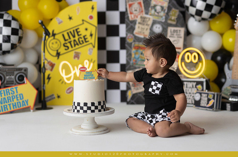One-year-old boy enjoying a Vans Warped Tour inspired first birthday milestone photography session, posing with a microphone, guitar, concert backdrop, checkerboard details, balloons, and rock concert props on a custom music-themed set at Studio 120 Photography in Ramona, serving San Diego and surrounding areas.