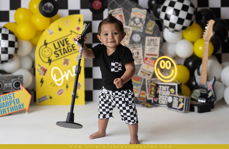 One-year-old boy enjoying a Vans Warped Tour inspired first birthday milestone photography session, posing with a microphone, guitar, concert backdrop, checkerboard details, balloons, and rock concert props on a custom music-themed set at Studio 120 Photography in Ramona, serving San Diego and surrounding areas.