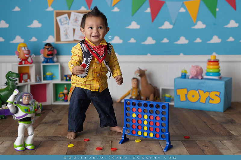 One-year-old boy dressed as Woody from Toy Story during a colorful milestone photography session, playing with classic toys including Rex the dinosaur, a cowboy hat, and ring toss games on a custom Toy Story inspired first birthday set at Studio 120 Photography in Ramona, serving San Diego and surrounding areas.