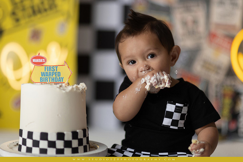 One-year-old boy enjoying a Vans Warped Tour inspired first birthday milestone photography session, posing with a microphone, guitar, concert backdrop, checkerboard details, balloons, and rock concert props on a custom music-themed set at Studio 120 Photography in Ramona, serving San Diego and surrounding areas.