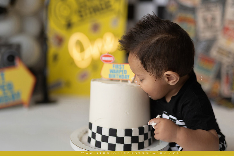One-year-old boy enjoying a Vans Warped Tour inspired first birthday milestone photography session, posing with a microphone, guitar, concert backdrop, checkerboard details, balloons, and rock concert props on a custom music-themed set at Studio 120 Photography in Ramona, serving San Diego and surrounding areas.