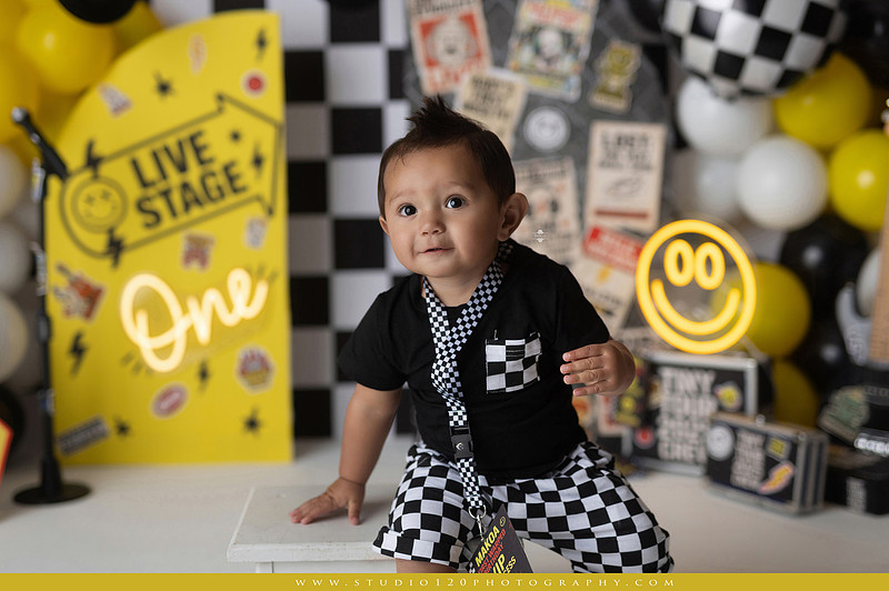 One-year-old boy enjoying a Vans Warped Tour inspired first birthday milestone photography session, posing with a microphone, guitar, concert backdrop, checkerboard details, balloons, and rock concert props on a custom music-themed set at Studio 120 Photography in Ramona, serving San Diego and surrounding areas.