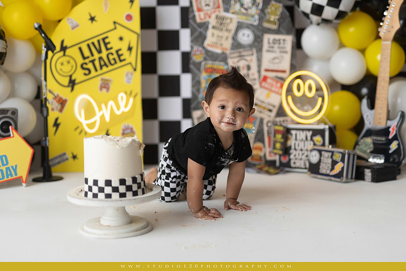 One-year-old boy enjoying a Vans Warped Tour inspired first birthday milestone photography session, posing with a microphone, guitar, concert backdrop, checkerboard details, balloons, and rock concert props on a custom music-themed set at Studio 120 Photography in Ramona, serving San Diego and surrounding areas.