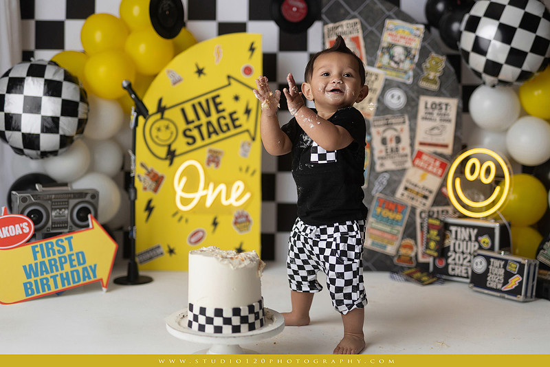 One-year-old boy enjoying a Vans Warped Tour inspired first birthday milestone photography session, posing with a microphone, guitar, concert backdrop, checkerboard details, balloons, and rock concert props on a custom music-themed set at Studio 120 Photography in Ramona, serving San Diego and surrounding areas.