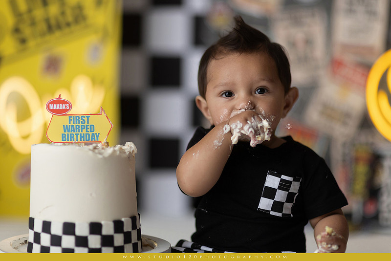 One-year-old boy enjoying a Vans Warped Tour inspired first birthday milestone photography session, posing with a microphone, guitar, concert backdrop, checkerboard details, balloons, and rock concert props on a custom music-themed set at Studio 120 Photography in Ramona, serving San Diego and surrounding areas.