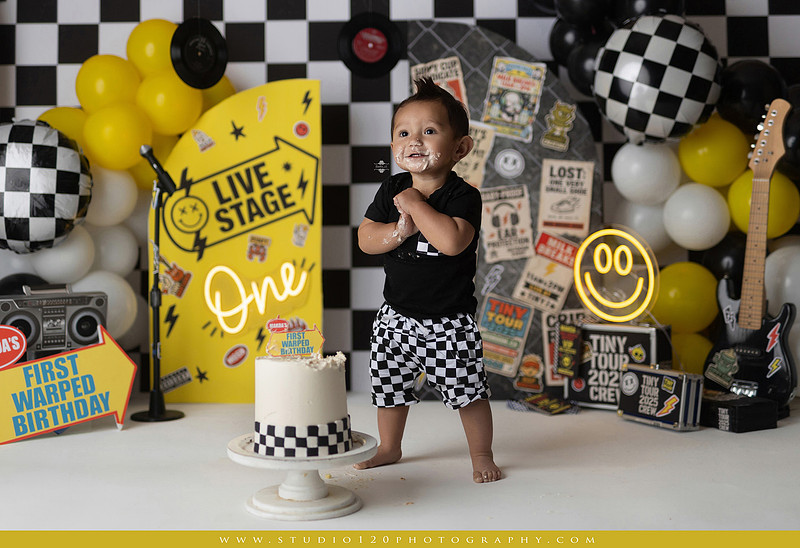 One-year-old boy enjoying a Vans Warped Tour inspired first birthday milestone photography session, posing with a microphone, guitar, concert backdrop, checkerboard details, balloons, and rock concert props on a custom music-themed set at Studio 120 Photography in Ramona, serving San Diego and surrounding areas.