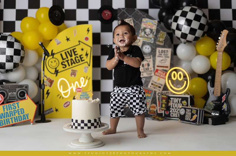 One-year-old boy enjoying a Vans Warped Tour inspired first birthday milestone photography session, posing with a microphone, guitar, concert backdrop, checkerboard details, balloons, and rock concert props on a custom music-themed set at Studio 120 Photography in Ramona, serving San Diego and surrounding areas.