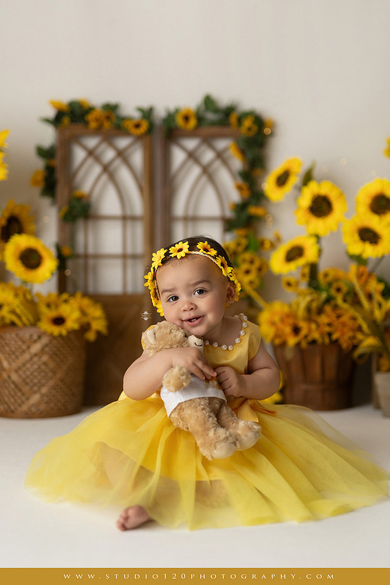 First birthday sunflower cake smash photography session featuring a baby girl in a yellow dress and sunflower headband, surrounded by bright sunflowers, rustic wood details, a teddy bear, and a yellow and white birthday cake at Studio 120 Photography in Ramona, California.
